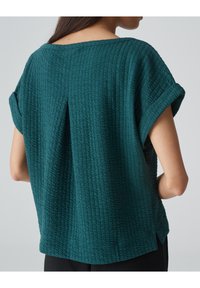 Vrouw in een tealgekleurde, getextureerde, kortmouwige top met opgerolde mouwen, gezien vanaf de achterkant tegen een effen achtergrond.