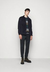 Μπουφάν από σουέντ ναυτικού, πουλόβερ πλεκτό με σχέδιο σε πράσινους και καφέ τόνους, σκούρο παντελόνι, καφέ μποτάκια μέχρι τον αστράγαλο. Το μοντέλο στέκεται μπροστά σε απλό φόντο.