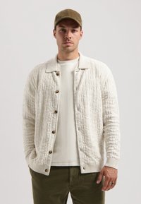 Texturirana bela pletena cardigan s ovratnikom, lesenimi gumbi, nosi se čez lahek pulover. Model nosi zeleni žametni klobuk in olivno zelene hlače.