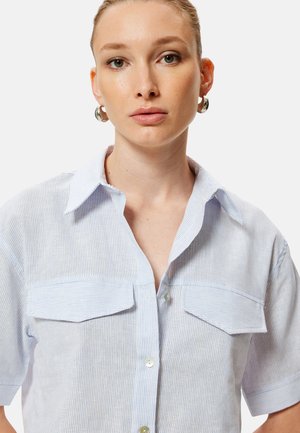 Jonge vrouw met lichtbruin haar naar achteren gebonden, draagt zilveren creolen en een lichtblauw gestreept kort mouwen overhemd met knopen.