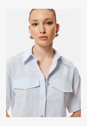 Jonge vrouw met lichtbruin haar naar achteren gebonden, draagt zilveren creolen en een lichtblauw gestreept kort mouwen overhemd met knopen.