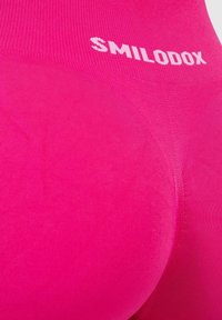 Fuksjowe legginsy wykonane z teksturowanej, elastycznej tkaniny, z bezszwowym wykończeniem i białym logo "SMILODOX" na pasku.