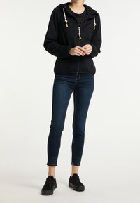 Femme portant un sweat à capuche noir à fermeture éclair avec des cordons blancs, un jean skinny foncé et des baskets noires, debout devant un fond uni.