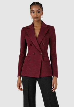 Donna che indossa un blazer doppiopetto aderente color bordeaux con dettagli a bottoni e pantaloni neri a gamba larga, in piedi davanti a uno sfondo semplice.