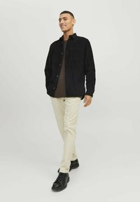 Zwarte corduroy overhemdjas met knopen aan de voorkant, twee borstzakken, gelaagd over een bruine gebreide trui, gecombineerd met crèmekleurige broek en zwarte sneakers.