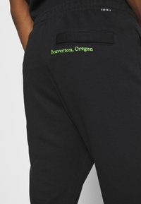 Svarta sweatpants med en texturerad tyg, elastisk midja och en bakficka med "Beaverton, Oregon" broderat i starkt grönt.