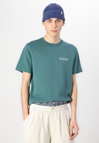 Camiseta de algodón color verde azulado con cuello redondo y mangas cortas que presenta el texto "Hotel de Palermo". Combinada con pantalones beige claros y un gorro azul de canalé.