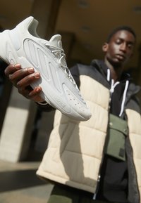 Valkoinen urheilukenkä, jossa on tekstuuri kuvioita, huomattavat nauhat ja pehmeä pohja, pidetään kädellä kaupunkitaustan edessä.