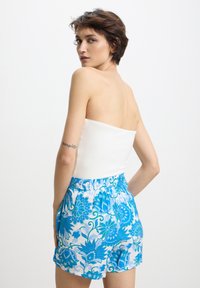 Donna con i capelli corti castani che indossa un top bianco senza spalline e pantaloni corti blu con stampa floreale, in piedi con le mani in tasca, che guarda oltre la spalla.