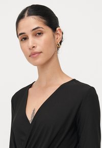 Vestito avvolgente nero con scollatura a V, realizzato in tessuto morbido. Presenta maniche corte e un tatuaggio sobrio visibile sulla parte superiore del petto.