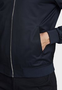 Chaqueta bomber azul marino de tela suave, con cremallera frontal, puños acanalados y bolsillos laterales. Detalle texturizado en la cinturilla.
