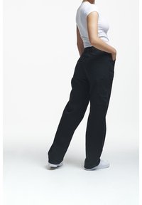 Donna che indossa pantaloni cargo neri a gamba larga, una maglietta bianca attillata a maniche corte e sneakers bianche, in piedi con le mani nelle tasche.