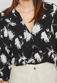 Blusa nera con motivo floreale bianco, dotata di scollatura a V profonda e maniche a tre quarti, abbinata a pantaloni chiari a vita alta.