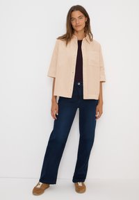 Camisa de botón beige claro con mangas cortas y un bolsillo en el pecho, combinada con unos vaqueros anchos azul oscuro y zapatillas marrones.