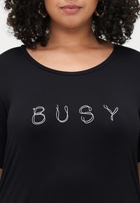 Svart T-skjorte med korte ermer og rund hals, med hvit tekst som leser "BUSY" i et lekent, loopet skrifttype på forsiden.