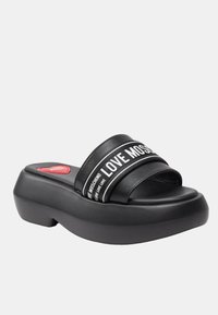 Sandali platform neri con due fasce larghe che presentano il testo "LOVE MOSCHINO" in bianco. Suola spessa e imbottita con un accento a forma di cuore rosso.