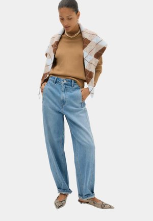 Donna con un maglione beige, jeans blu a vita alta, ballerine con stampa a serpente e un maglione a fantasia drappeggiato sulle spalle.