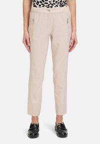 Beżowe spodnie o gładkiej fakturze, z bocznymi kieszeniami na zamek, z paskiem w talii z guzikem z przodu oraz zwężającą się nogawką. W połączeniu z czarnymi butami.