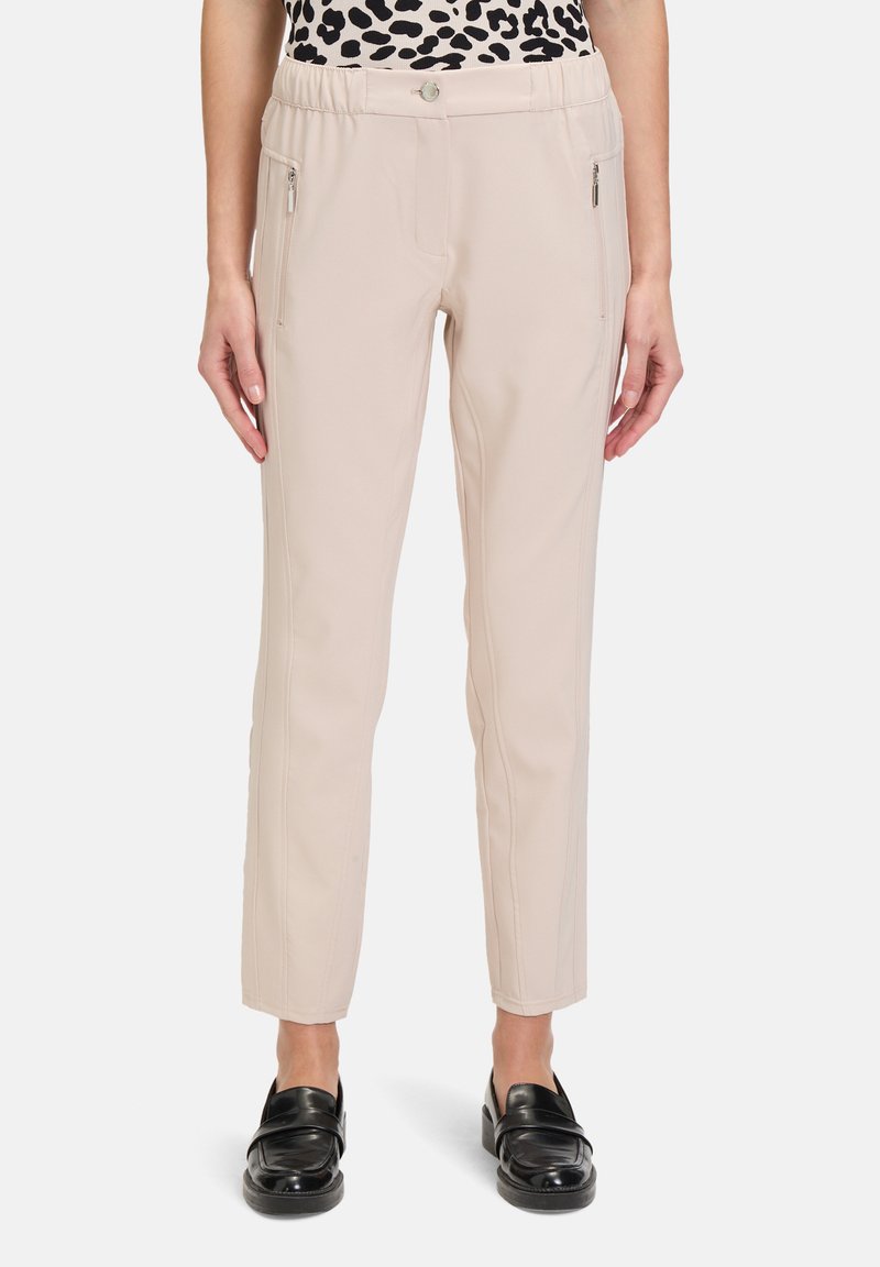 Beżowe spodnie o gładkiej fakturze, z bocznymi kieszeniami na zamek, z paskiem w talii z guzikem z przodu oraz zwężającą się nogawką. W połączeniu z czarnymi butami.