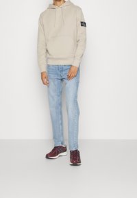 Sudadera con capucha de color beige claro, con bolsillo canguro y logotipo en la manga, combinada con jeans de lavado claro y zapatillas burdeos. Textura suave.