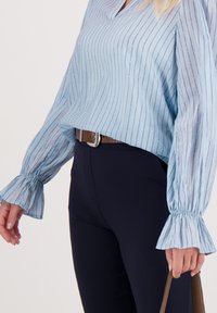 Lichtblauw gestreepte blouse met gerimpelde manchetten en subtiele glans, gecombineerd met donkere broek en een bruine riem, vastgehouden met een handtas.