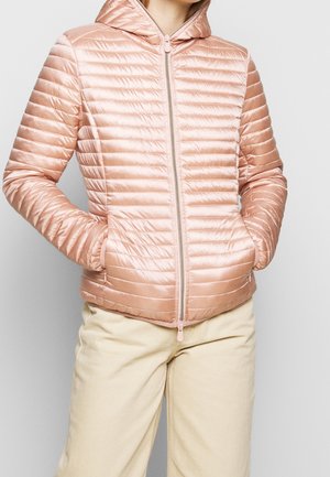 Kvinde iført en lyserød quiltet hættejakke med lynlås foran og hænder i lommerne, kombineret med lyse beige bukser.