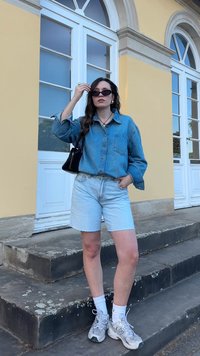 Jeans košulja na dugmad, svijetloplave kratke hlače, bijele čarape i sivi tenisice. Crna torba i sunčane naočale upotpunjuju odjeću. Stepenice su vidljive.