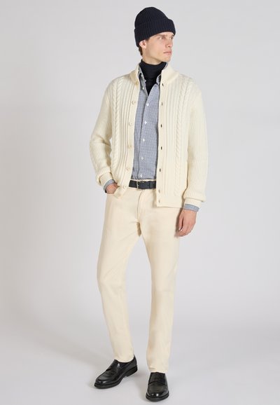Off-white kabelbrei cardigan, marineblauwe beanie, geruit overhemd, zwarte riem, crèmekleurige broek en zwarte instappers. Soepele textuur en casual pasvorm.