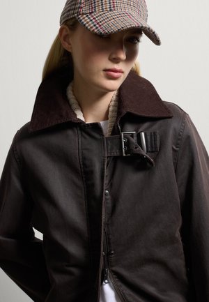 Bruine katoenen jas met een brede corduroy kraag, drukknoopsluiting en een gespdetail; gedragen met een geruite pet. Wol textuur zichtbaar rond de kraag.