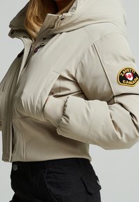 Veste beige zippée avec col montant et écusson Zavetti Canada sur la manche, portée avec une main dans la poche et un pantalon noir.