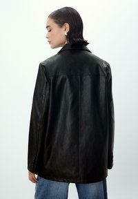 Giacca in pelle nera con una silhouette strutturata, rever a punta e polsini con bottoni. La texture liscia e la lucentezza sottile arricchiscono il design.