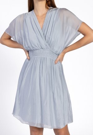 Kobieta ubrana w jasnoniebieską sukienkę do kolan z głębokim dekoltem w kształcie litery V i marszczonym pasem, z rękami opartymi na biodrach na białym tle.