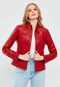 Rode leren jas met een ritssluiting aan de voorkant, twee borstzakken en zij-ritsen. Heeft een staande kraag en een aansluitend ontwerp, gecombineerd met lichtblauwe jeans.