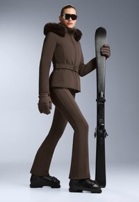 Hnědý lyžařský outfit s kapucí s kožešinovým lemem, fitted bunda a rozšířené kalhoty. Černé lyžařské boty a rukavice. Drží černou lyži.