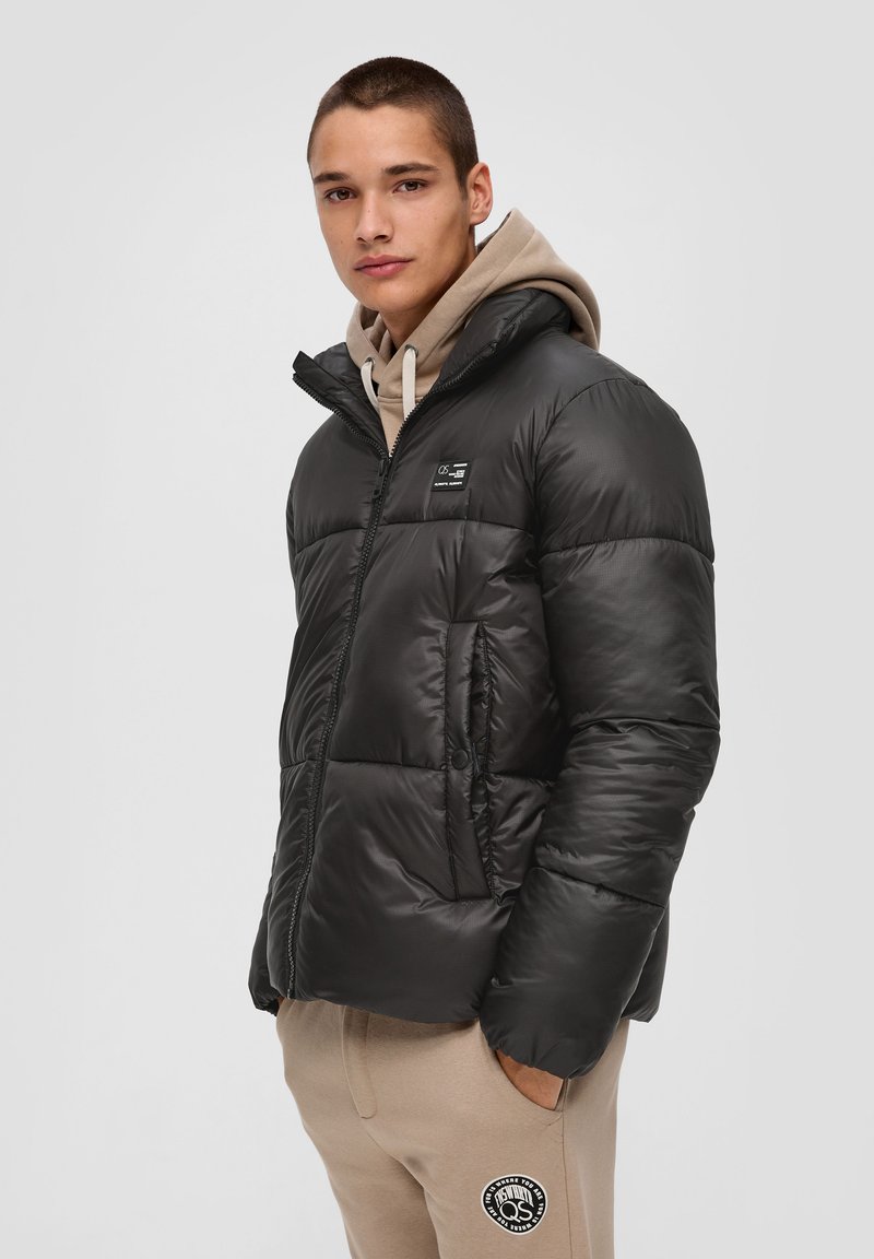 Schwarze Pufferjacke mit Reißverschluss vorne, hohem Kragen und einem kleinen Logo-Patch. Verfügt über ein gestepptes Design und zwei seitliche Taschen.