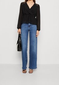 Blusa negra con detalles de encaje, combinada con jeans azul de tiro alto y pierna ancha. La modelo lleva un bolso negro brillante y sandalias negras con tiras.