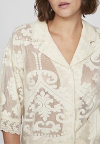 Camicia in pizzo color bianco sporco con motivo floreale, colletto, maniche corte e bordi decorativi in pizzo ai polsini. Chiusura frontale con bottoni.