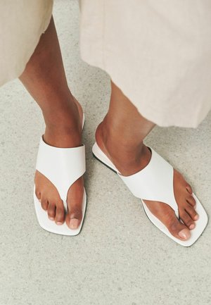 Lábak fehér bőrből készült, könnyen felvehető szandálban, széles pántokkal és négyzet alakú orr-résszel világos texturált padlón, részben látható bézs ruhadarab mellett.