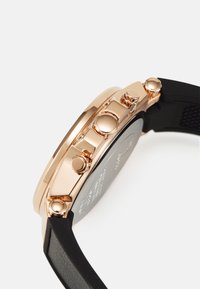 Montre en or rose avec un bracelet en silicone noir, couronne texturée et trois poussoirs. Elle possède un boîtier rond et un fond de boîtier noir.