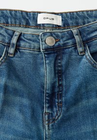 Blauwe spijkerbroek van denim met bruine stiksels, metalen knoop met gravure "OPUS" en wit label met zwarte "OPUS" tekst op de tailleband.