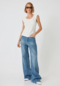 Witte ribgebreide tanktop met ruche mouwen, gecombineerd met hoge taille, wijde spijkerbroeken in lichtblauw en witte puntige schoenen.