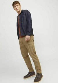 Denimjack met knoopsluiting, bruine shirt eronder, cargobroek in khaki en donkere laarzen met veters en lichte zolen.