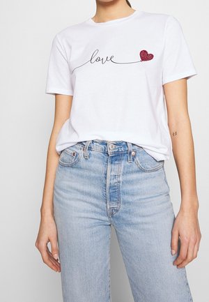 Kobieta w białej koszulce z napisem „love” w czarnym stylu pisma i czerwonym sercem z koralików, w połączeniu z jasnoniebieskimi, taliowanymi dżinsami.