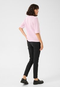 Heledad roosa, lühikeste varrukatega kootud topp avatud kudumisega; sobitatud mustade kitsaste pükste ja mustade kingadega, vaade tagant.