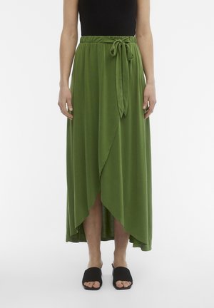 Vrouw draagt een groene overslagrok met een hoge voorzijde en lage achterkant, met een gestrikte strik in de taille en zwarte open-toe instapsandalen.