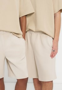 Δύο άτομα που φοράνε μπεζ oversized T-shirts και ταιριαστά μπεζ σορτς με τσέπες, στέκονται δίπλα-δίπλα σε λευκό φόντο.