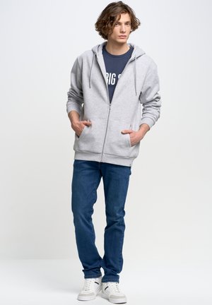 Grijze zip-up hoodie met een zachte textuur, voorzien van een trekkoord in de capuchon en voorzakken. Draag het over een marineblauw shirt en gecombineerd met blauwe jeans.