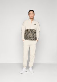 Beige Reißverschluss-Fleece-Sweatshirt mit einem mittleren Abschnitt im Leopardenmuster, kombiniert mit beigen Jogginghosen und weißen Sportschuhen. Schwarzes Logo sichtbar.