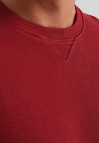 Mörkröd sweatshirt med rundad halsringning, texturerad tyg och ribbade ärmslut. Har en subtil V-söm detalj vid kragen.