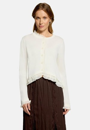 Woman with light brown hair wearing a white buttoned cardigan with ruffles over a long brown skirt with lace details.